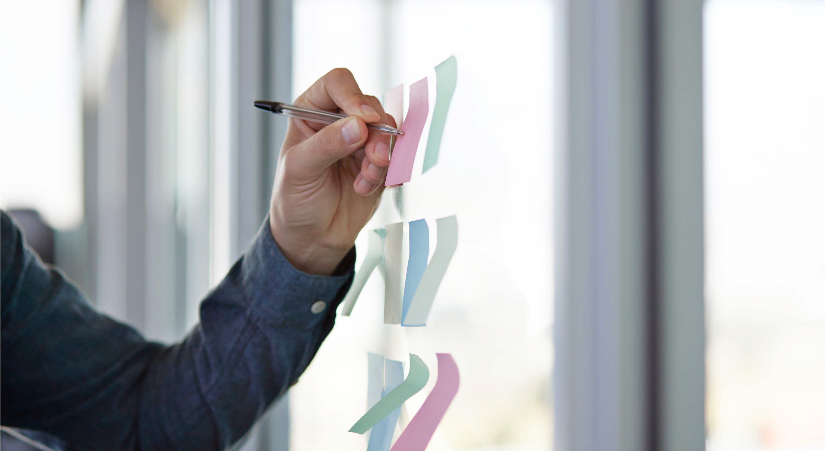 A person’s left arm writes on sticky notes that are placed on a glass wall.