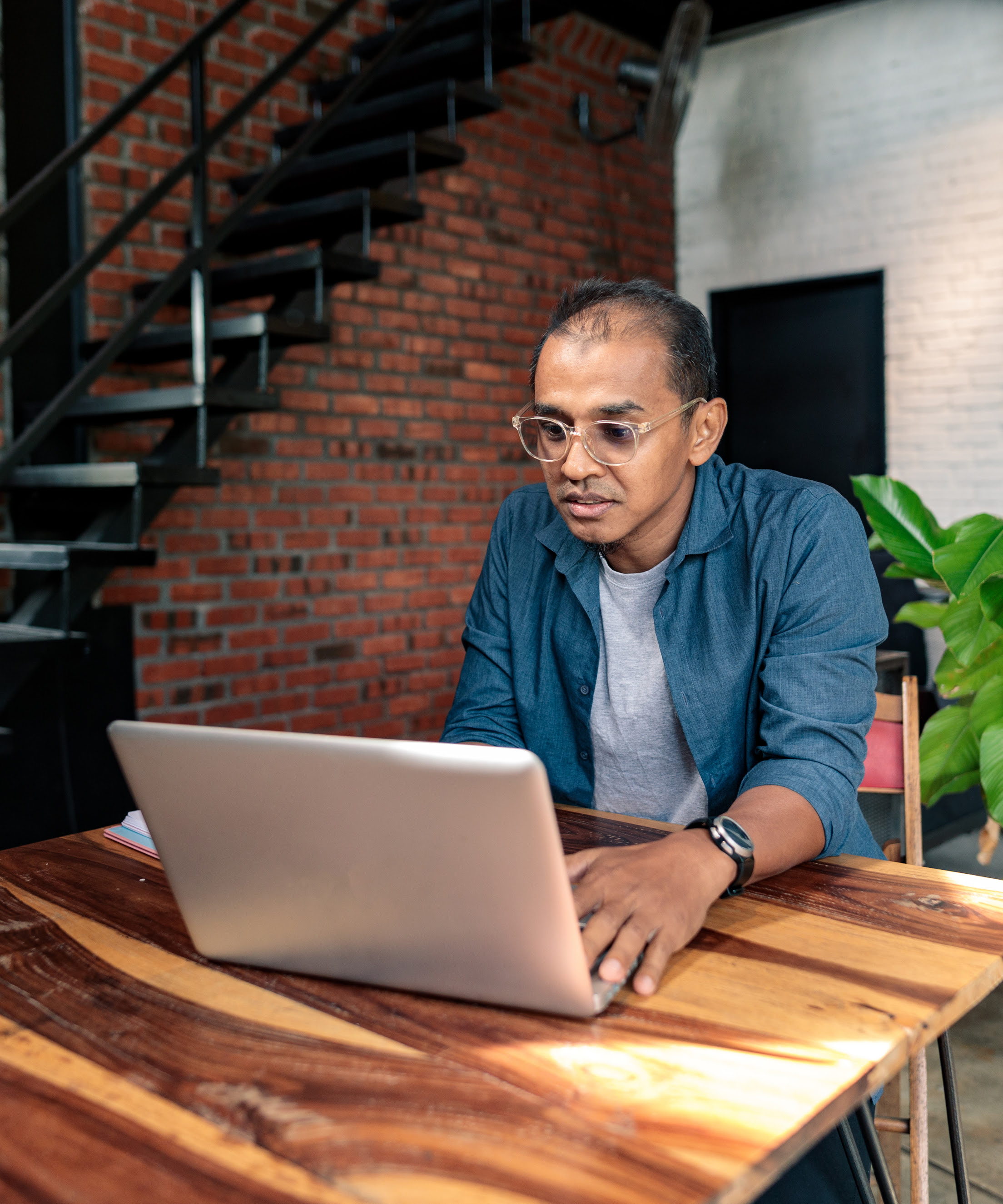 A man with glasses sits at a high top wooden table while working on his laptop.