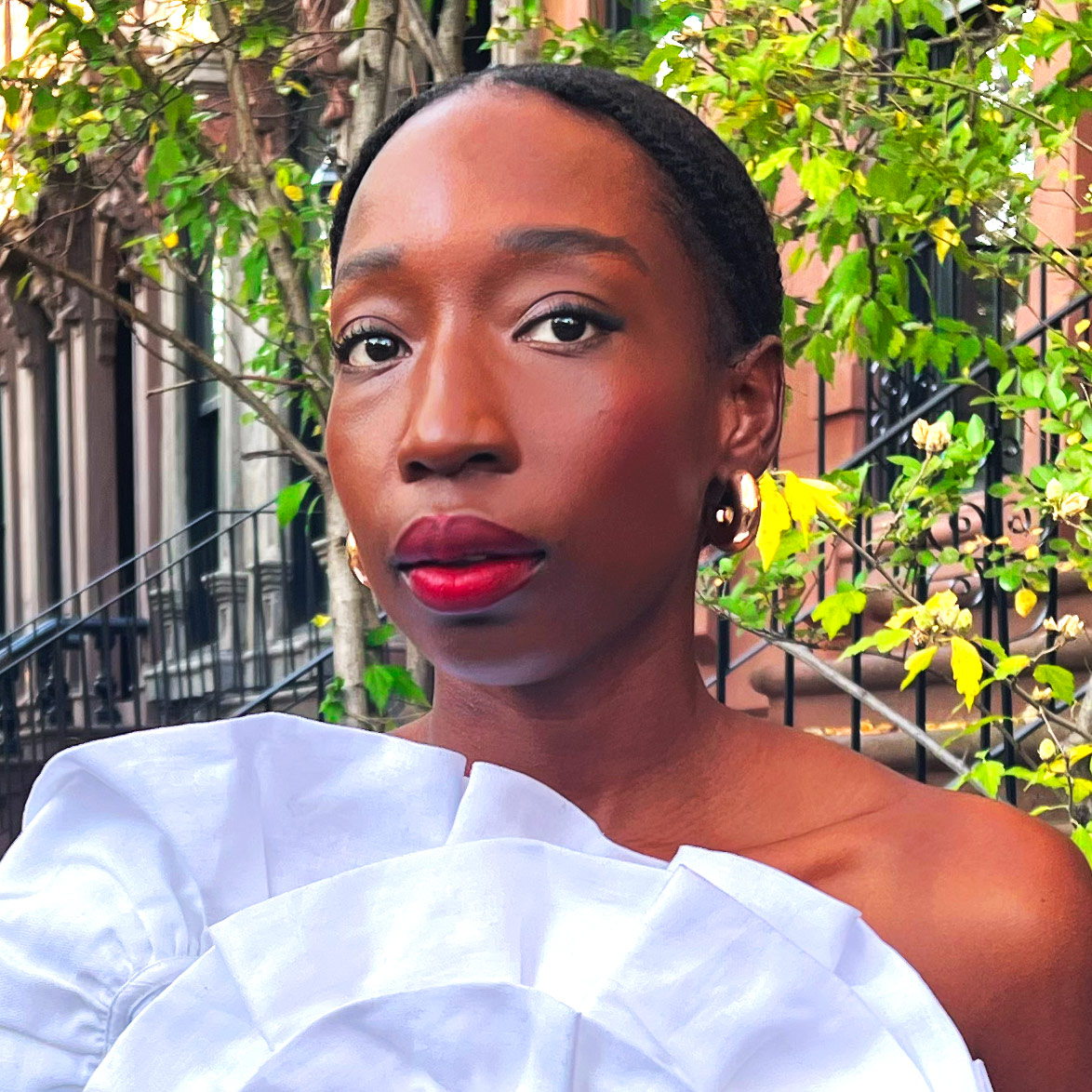 Close shot of a woman's face. She has black hair is wearing a white top and gold earrings. She is standing in front of a brownstone houses and green trees.