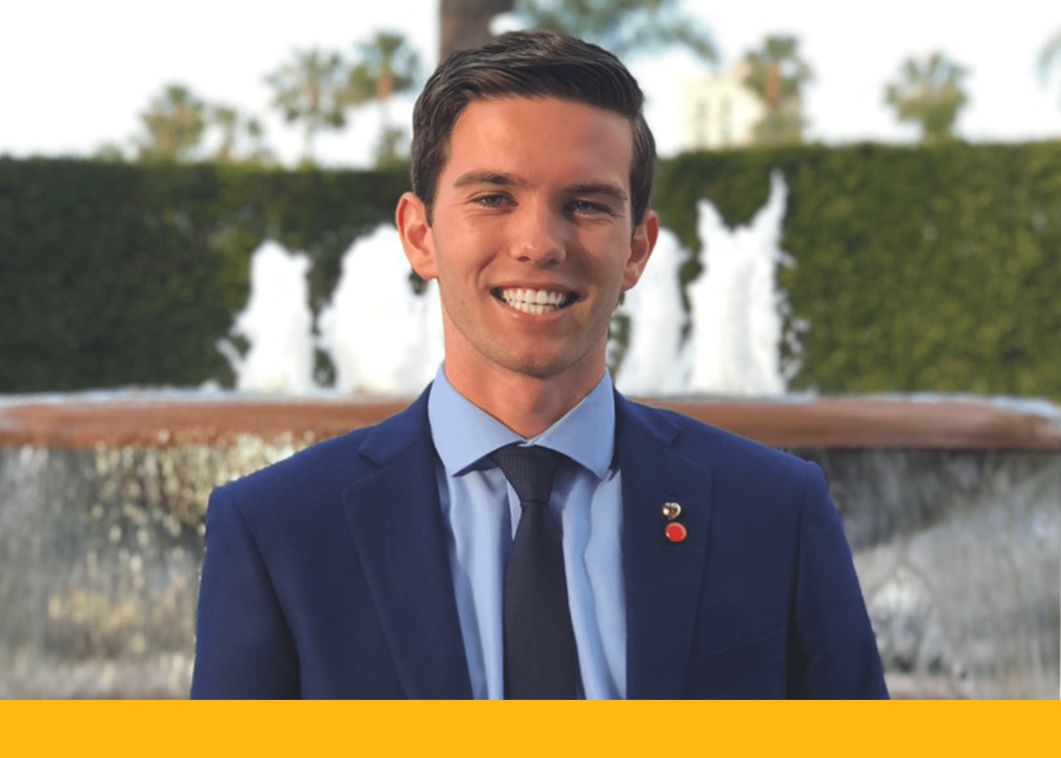 student in a suit smiling at the camera with a fountain in the backdrop