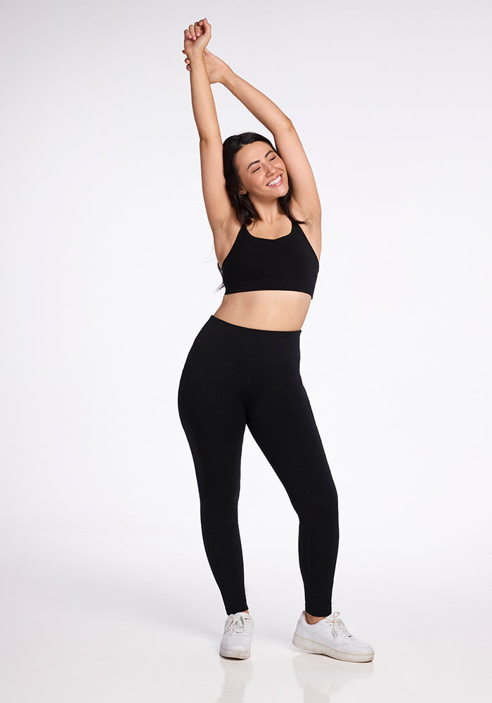 A woman stands with her arms stretched above her head, smiling and wearing a black sports bra, Frost Duralite� Leggings by Woolx in black, and white sneakers. The background is plain white.