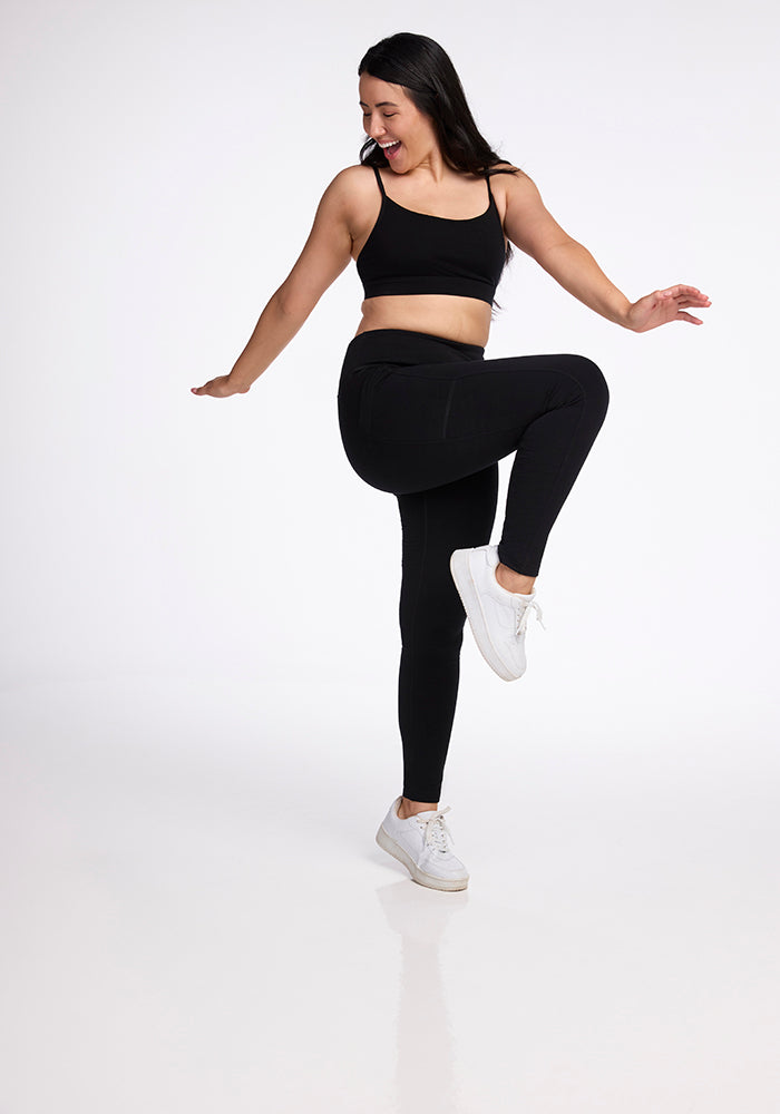 A woman wearing Woolx's Frost Duralite� Leggings in black, featuring convenient side pockets, and a pair of white sneakers is smiling as she balances on one leg with her arms outstretched. She is posing against a plain white background.