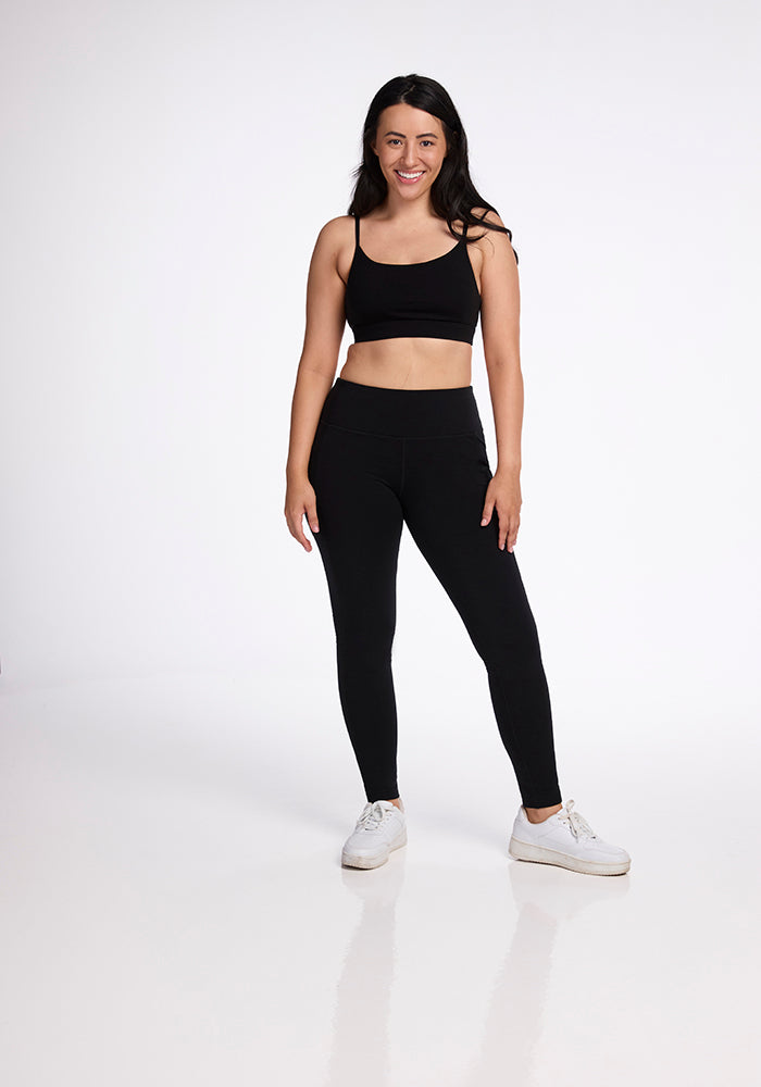 A woman with long dark hair is smiling and standing in a white space, wearing a black sports bra, Woolx Frost Duralite� Leggings - Black with side pockets, and white sneakers. She has her arms relaxed by her sides, and her reflection is faintly visible on the shiny floor.