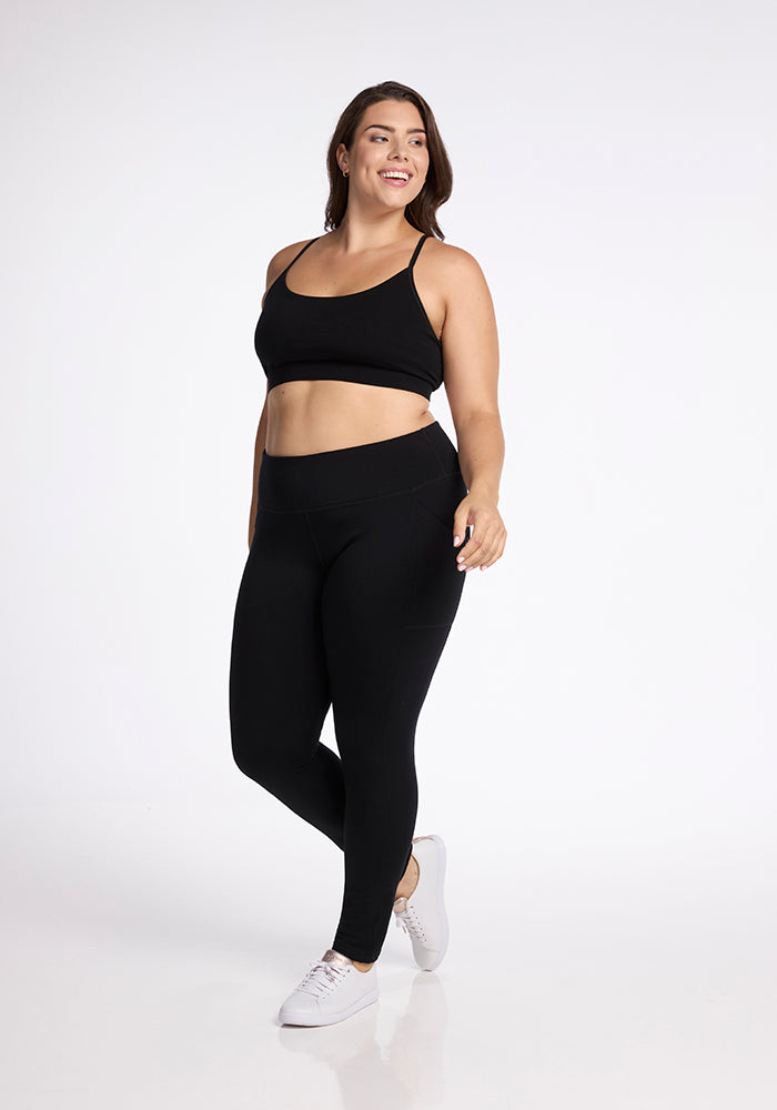 A woman poses confidently in Woolx's black Frost Duralite� Leggings and a matching sports bra against a simple white background. She has brown hair, wears white sneakers, and smiles while looking slightly to the side.