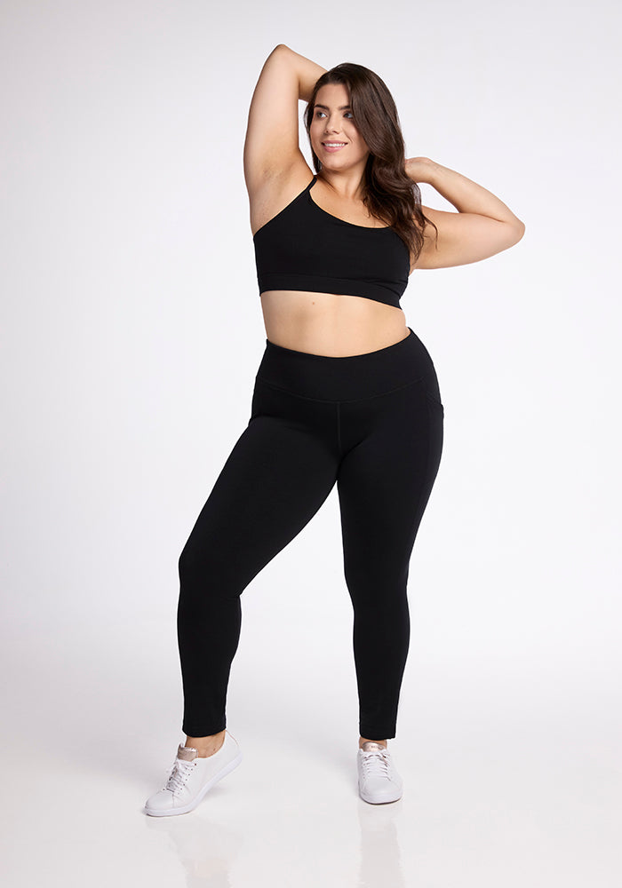 A woman with long brown hair poses confidently against a white background. She is dressed in a black sports bra, Woolx Frost Duralite� Leggings in Black, and white sneakers. Her left arm is bent with her hand resting on the back of her head, and her right arm is relaxed by her side.