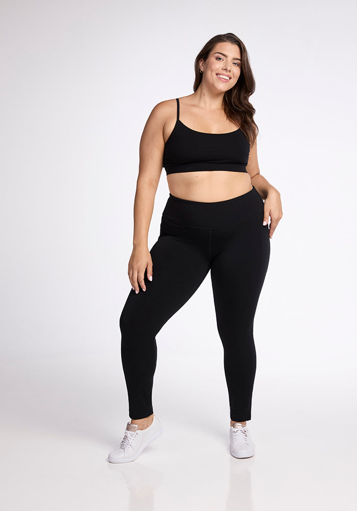 A woman poses confidently in Woolx's black Frost Duralite� Leggings and a matching sports bra, paired with white sneakers. She smiles and stands with one hand on her hip against a plain white background.