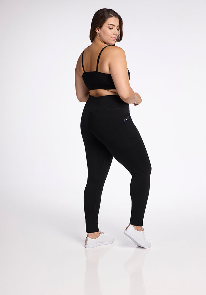 A woman stands facing away, slightly turning to her left. She is wearing a black sports bra, Frost Duralite� Leggings - Black by Woolx, and white sneakers. The background is plain white, highlighting her athletic outfit.