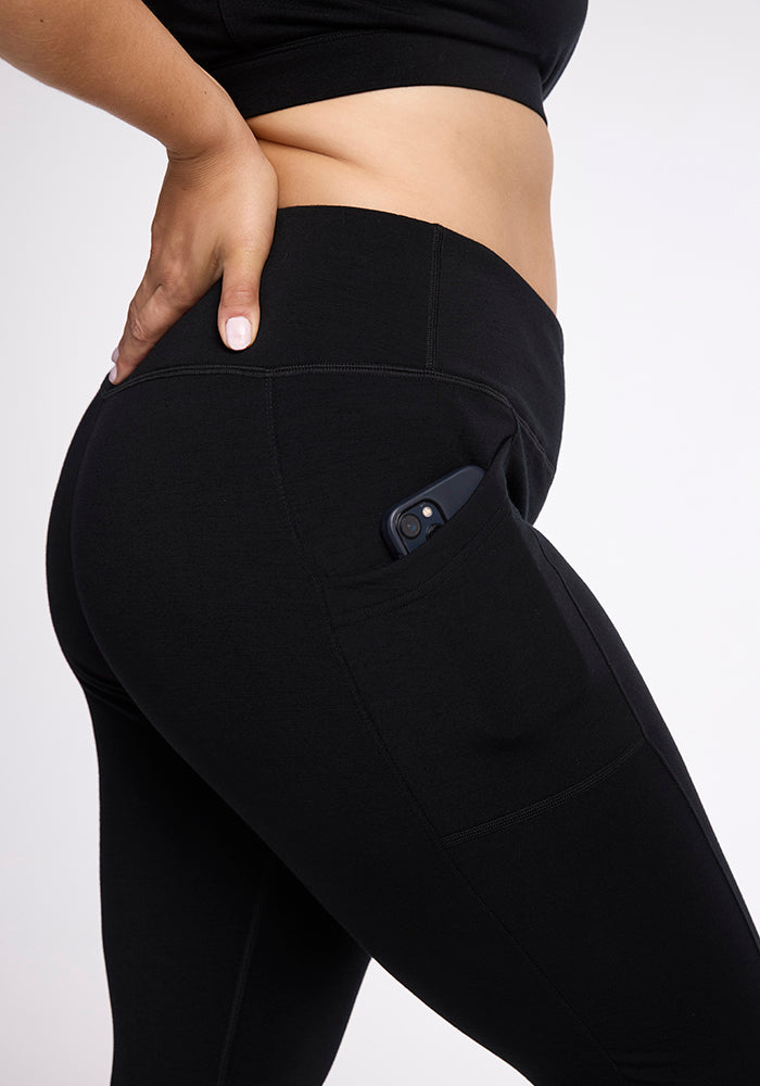 Close-up of a person's torso and upper leg, showing them wearing black Frost Duralite� Leggings from Woolx with a phone partially tucked into a side pocket. The person is placing their hand on their hip, and they are dressed in a matching black top.