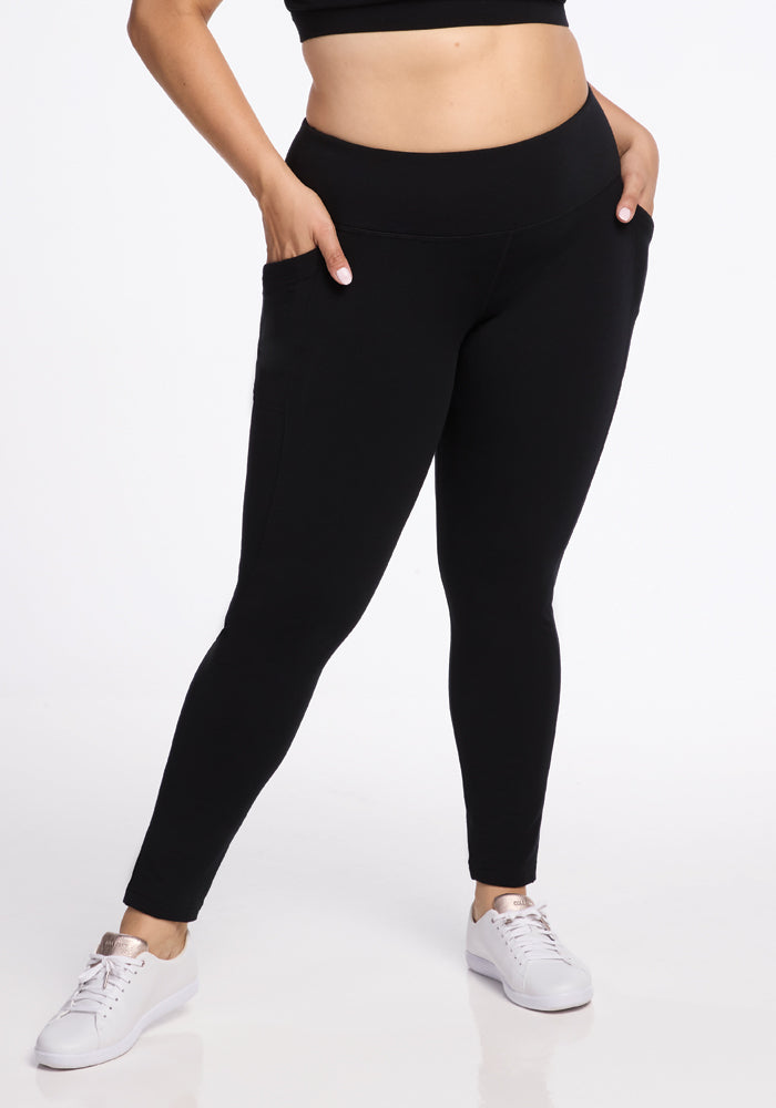 A person is wearing Woolx Frost Duralite� Leggings in black, with their hands in the side pockets. They are also sporting a black crop top and white sneakers. These warm leggings function as the perfect baselayer for any outfit. The background is plain white. 