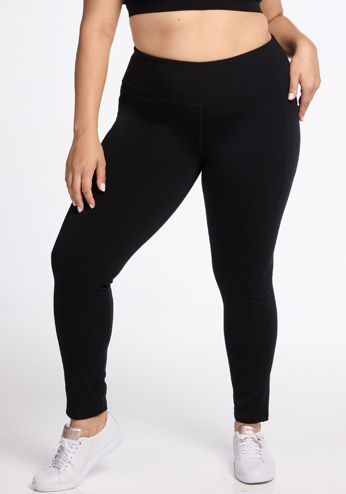 A person with light skin is wearing Woolx's Frost Duralite� Leggings in black paired with a black crop top. They are also wearing white sneakers against a plain white backdrop. The individual stands with one arm hanging loose while the other rests on their hip. 