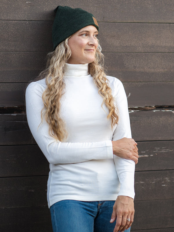 Woman wearing buttercream peyton turtleneck and a pine baylor beanie against wall