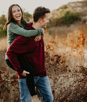 Couple standing in field wearing woolx. The man is carrying the woman on his back and laughing. 