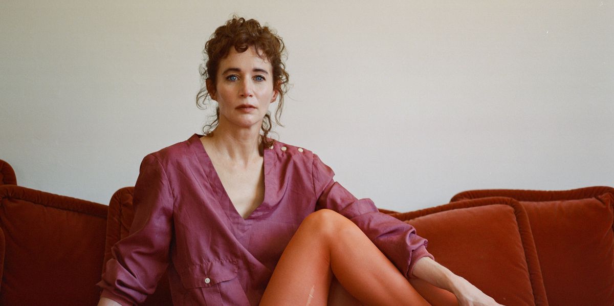 Miranda July sitzt auf einem roten Sofa in Los Angeles, April 2024, im Hintergrund ein Kunstwerk an der Wand. Sie tr�gt ein violettes Oberteil und orangefarbene Strumpfhosen.