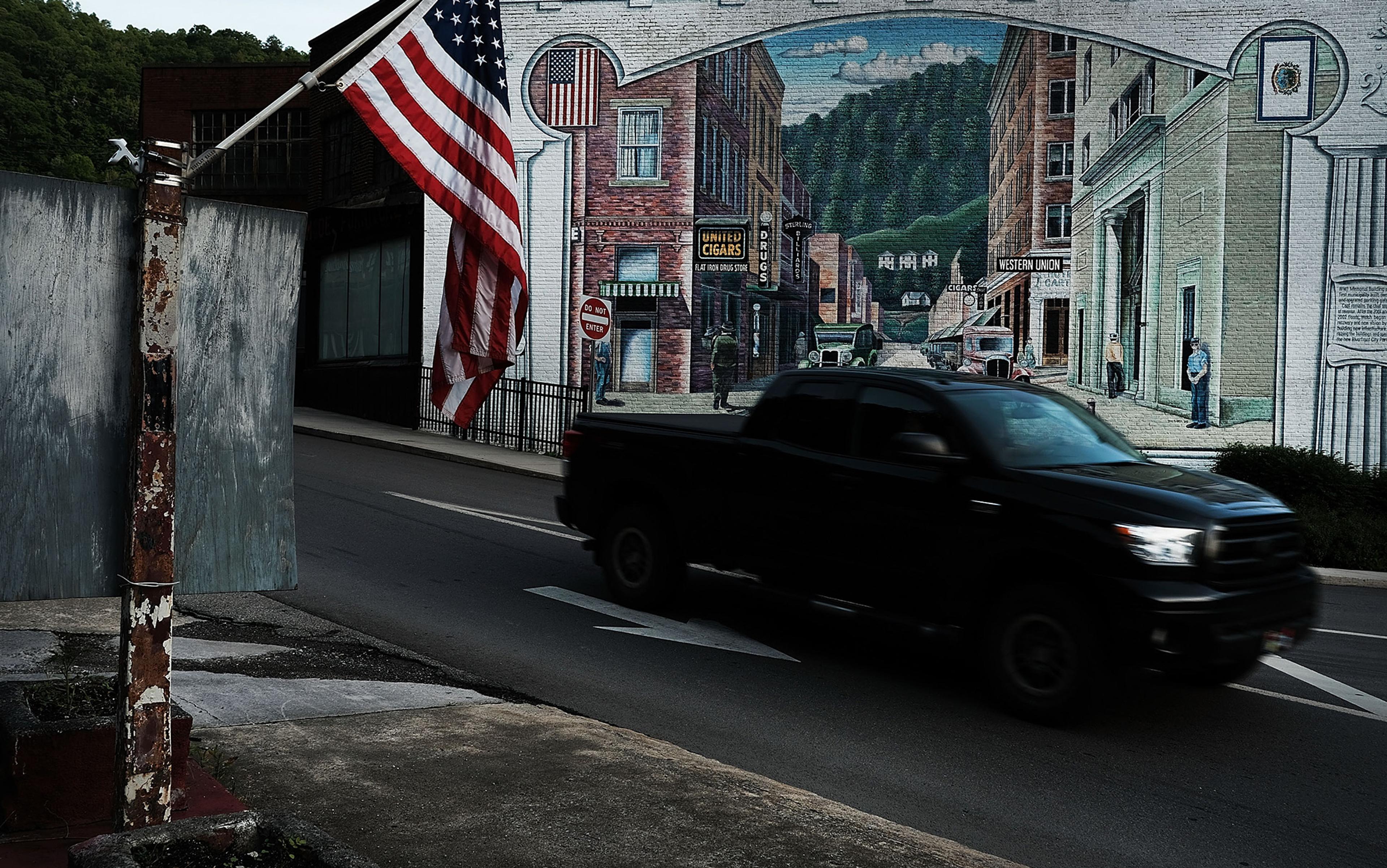 A street with an American flag a mural of a vintage town scene and a black pickup truck passing by.