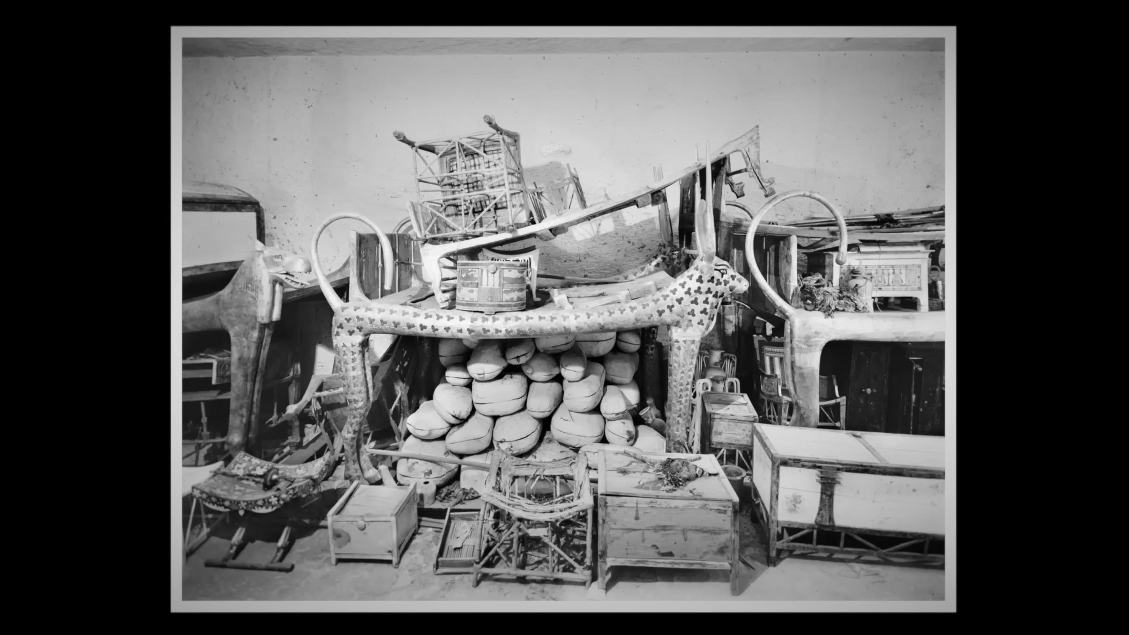 Black and white photo of ancient Egyptian artefacts in storage, including statues, chests, chairs, and bundles, within a room.