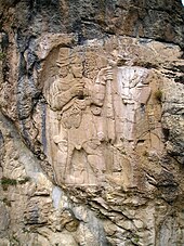 La photographie montre un rocher sculpté ; le bas-relief apparait en gris-beige sur une roche gris-bleu maculée de trainées gris-sombre. Le roi hittite est à gauche. Il est barbu, et tient un rameau portant deux grappes de raisin. Face à lui, le dieu a perdu sa tête et ses pieds à cause des outrages du temps