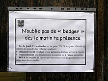 Gros plan d’une affichette de format A4 horizontal glissée dans une chemise plastique et agrafée à un panneau de bois. Dans un encadré gras, et sous le titre « N’oublie pas de « badger » dès le matin ta présence », l’avis annonce : « Dès le jeudi 21 septembre et ce pour tous les jours d’école (y compris le samedi), les enfants viendront : - obligatoirement « badger » leur présence dès leur arrivée à l’école, - éventuellement leur inscription à la cantine ou à l’étude surveillée. Tous les enfants de l’école devront donc avoir en permanence leur badge dans leur cartable. »