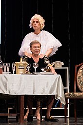 Photographie d'une scène de théâtre. Un homme blanc en robe noire est assis derrière une table bourgeoise tandis qu'un autre, en perruque blonde bon marchée et haut blanc en tule, lui masse les épaules.