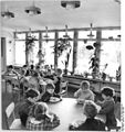 Dans une salle lumineuse dont l’un des côtés est vitré et décoré de plantes en pots disposés sur l’appui de fenêtre intérieur, des enfants de maternelle sont assis, par groupes de 6 ou 7, à des tables rectangulaires ou rondes, et mangent à la cuiller le repas servi dans des assiettes de faïence.