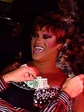 Photographie d'une drag queen Noire aux cheveux courts. Des billets de dollar sont glissés dans son décolté, que de mains blanches, visibles sur la photo, viennent de déposer.