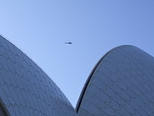 Top of an unrecognizable curvy building under blue sky with a helicopter so far in the distance that it looks like a gnat