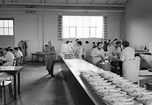 Photo en noir et blanc de l’extrémité d’une grande salle. À droite, sur un comptoir, une vingtaine de plats partiellement servis attendent d’être complétés par des femmes en tablier blanc. Au centre, un homme moustachu, en tablier de travail, s’avance parallèlement au comptoir et semble rouler une cigarette. À gauche, quelques hommes attablés mangent et discutent.