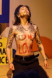 Drag king à genoux, yeux fermés, torse nu, sur lequel est inscrit en grandes lettres rouges "it's not love". Il porte une large ceinture noire et un pantalon de costume noir.