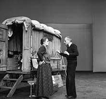 Photographie en noir et blanc. Devant une roulotte en bois, bâchée, porte ouverte, à l’intérieur de laquelle on accède par un escalier de deux marches, le metteur en scène donne des indications à l'actrice. Tous deux sont de profil, lui en vêtement de ville (pantalon et pull d’où dépasse le col clair d’une chemise), elle en costume de scène : cheveux serrés en petit chignon sur la nuque, corsage à longues manches sombres recouvert d’une casaque plus claire serrée à la taille par une ceinture qui retient une sacoche que l’on devine portée au côté gauche, et longue jupe sombre à petits motifs clairs répétitifs.