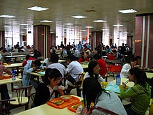 Dans une très grande salle d’architecture moderne, parsemée de massifs piliers carrés bordeaux et blancs, des étudiants sont assis à des tables pour 4 personnes et y mangent ou y discutent. Dans un angle de la salle, une TV est allumée.