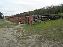 Ce long bâtiment d’un seul étage, au toit plat, semble être enfoncé dans la pente d’un terrain herbeux que clôture un bois de feuillus. La façade en briques est percée de deux portes et de six petites fenêtres.