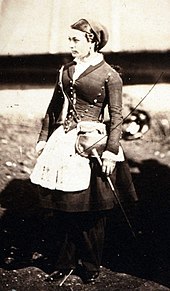 Portrait photographique sépia d’une jolie femme en uniforme, prise de trois-quart. La cantinière porte une jupe ballonnée au-dessus d’un pantalon long. Sur la jupe un tablier blanc. Le haut de l’uniforme est constitué d’une veste cintrée, moulante, à longues manches, boutonnée à l’avant et se terminant en pointe qui repose sur le tablier ; l’échancrure en V du col laisse apercevoir la chemise blanche soigneusement fermée jusqu’au cou ; la chemise dépasse aussi des manches jusqu’à reposer sur le dos de la main. Le marquage de la taille fine est accentué par une décoration de boutons qui part de chaque épaule et descend vers la pointe de la veste. La femme, portant une coiffe qui emprisonne les cheveux, porte une sacoche en bandoulière et tient à la main une baguette.