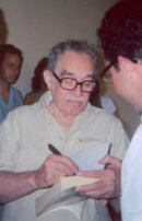 Grey haired man with a moustache and glasses stands while autographing books.