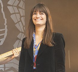 Femme jeune avec les cheveux longs châtains, photographiée de face, souriante ; le manche d'une basse et la main du bassiste est visible dans l'arrière-plan à gauche.