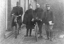 Photographie noir et blanc de quatre hommes en uniforme, deux sont sur des bicyclettes.