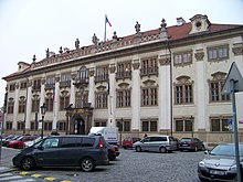 Barevná fotografie s pohledem na honosný zdobený dům (Nostický palác), v přední části parkoviště a řada zaparkovaných aut