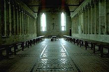 Le long des murs rythmés par des colonnades, d’une longue et haute salle, chichement éclairée par deux hautes baies deux rangées de tablées et bancs se font face. Au fond, sous une grande croix, se trouve placée perpendiculairement la table réservée aux supérieurs.