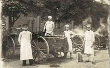 Des soldats allemands, en tabliers blancs et toques, posent pour le photographe autour de charrettes munies d’énormes roues ; de la vapeur s’élève de la seconde charrette.