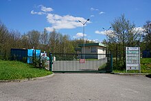 Photo couleur d'une route avec en arrière plan l'entrée grillagée d'une déchèterie aux bennes bleu