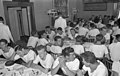 Dans une salle décorée de tableaux, des rangées de longues tables et chaises où sont assis de jeunes marins en T-shirts blancs qui viennent de terminer leur gâteau de dessert et leur café. Certains fument une cigarette. Deux serveurs sont en pantalon sombre et veste blanche.