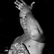 Photographie en noir et blanc d'une drag queen noire. Elle porte un bustier qui laisse apparaître son torse et du maquillage sur les yeux.