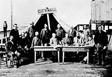 Men standing and sitting around two tables, facing the c�mera. A large tent behind them has a wooden sign that reads "City Hall"