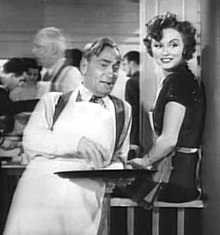 Photo en noir et blanc. William Demarest, en tablier blanc de serveur et tenant un plateau à la main s’appuie du dos contre un comptoir auquel est assise Cheryl Walker ; tous deux regardent, amusés, dans la direction du photographe.