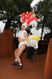 Photographie d'une sculpture de cire. Dans un décor qui rappelle le désert australien, une drag queen blanche porte une tenue de caranaval comportant notamment de longues plumes sur le dos.