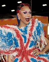 Photographie d'une drag queen noire souriante. Ses cheveux teints en rouge sont plaqués au gel et elle porte une robe aux couleurs du drapeau britannique.