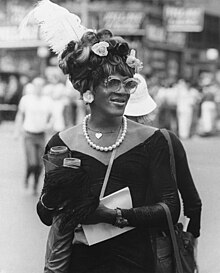 Photographie en noire et blanc d'une drag queen et femme trans noire. Elle porte une longue robe de velour foncé ainsi qu'un colier de perle. Sa tenue est élégante et raffinée.